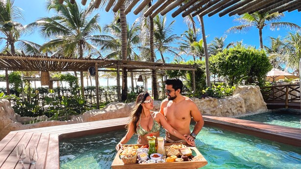 Ein Paar genießt eine Mahlzeit am Pool mit einer Holzterrasse und Palmen im Hintergrund.