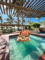 A couple enjoying a meal by a pool with a wooden deck and palm trees in the background.