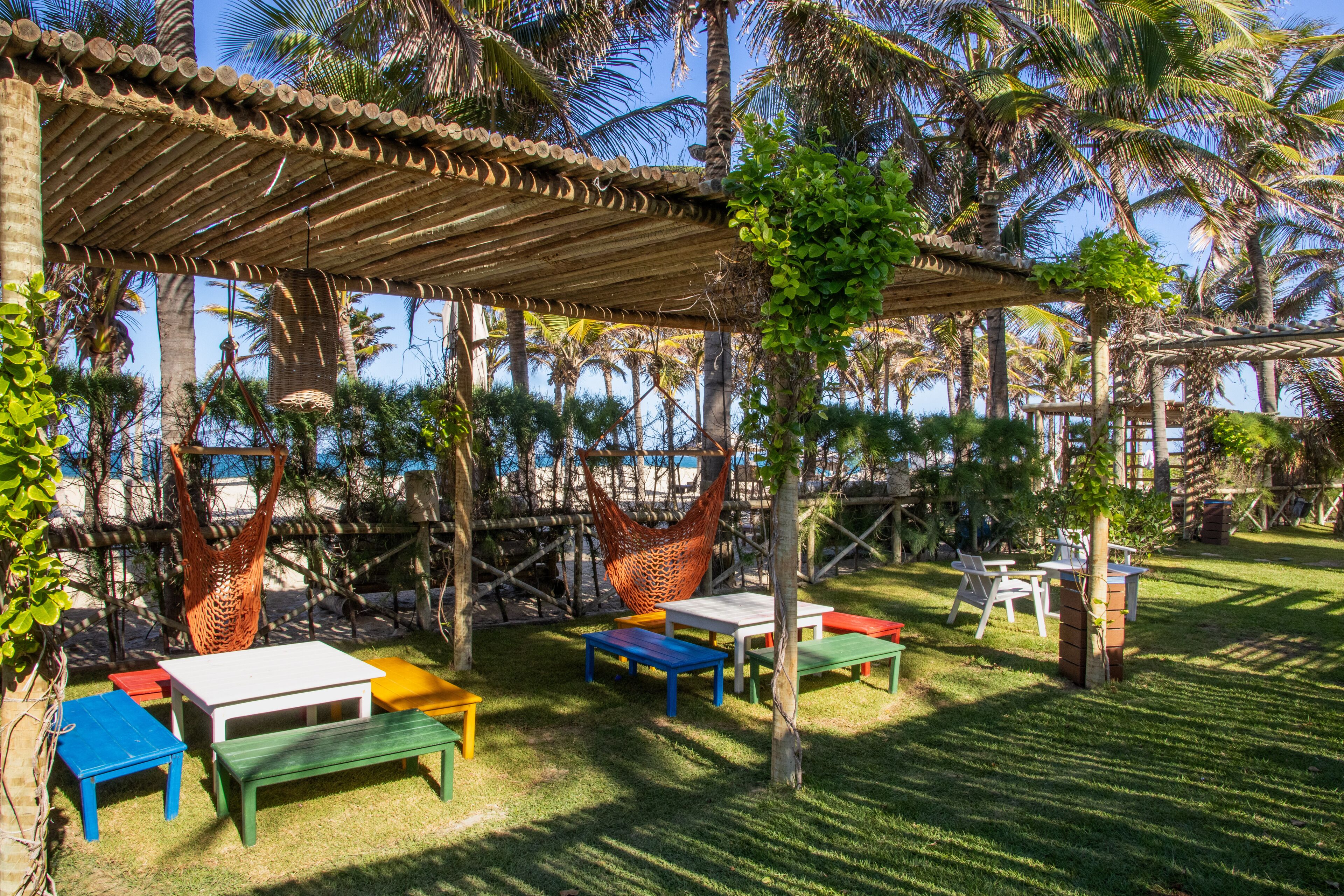 A covered outdoor seating area with colorful tables and benches, surrounded by palm trees and a clear sky.