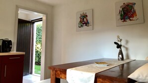 Ein Zimmer mit einem Holztisch, einer weißen Wand mit zwei gerahmten Kunstwerken, einer kleinen Vase mit Blumen und Blick auf Grünfläche draußen.