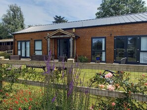 Une maison en bois à deux étages, dotée d’un porche à l’avant, de grandes fenêtres et d’un jardin clôturé où poussent diverses plantes et fleurs.