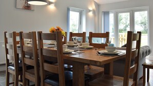 Zona pranzo con tavolo in legno apparecchiata per quattro persone, circondata da sedie in legno. Due lampade a sospensione illuminano il tavolo, e una finestra offre vista su piante verdi.