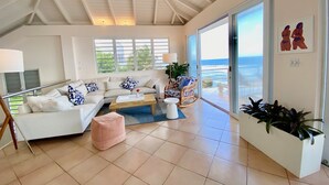 Uma sala de estar espaçosa com teto alto, uma grande janela com vista para o oceano, um aconchegante conjunto de sofás e uma área de jantar com mesa e cadeiras.