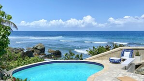 Piscina com borda curva, rodeada por um deck de pedra e área de estar com vista para o oceano.