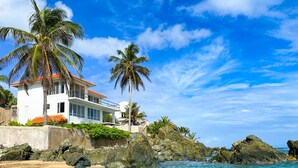 Uma casa costeira com fachada branca, telhado de telhas vermelhas e várias varandas. Está localizada numa costa rochosa com águas azul-claras e palmeiras.