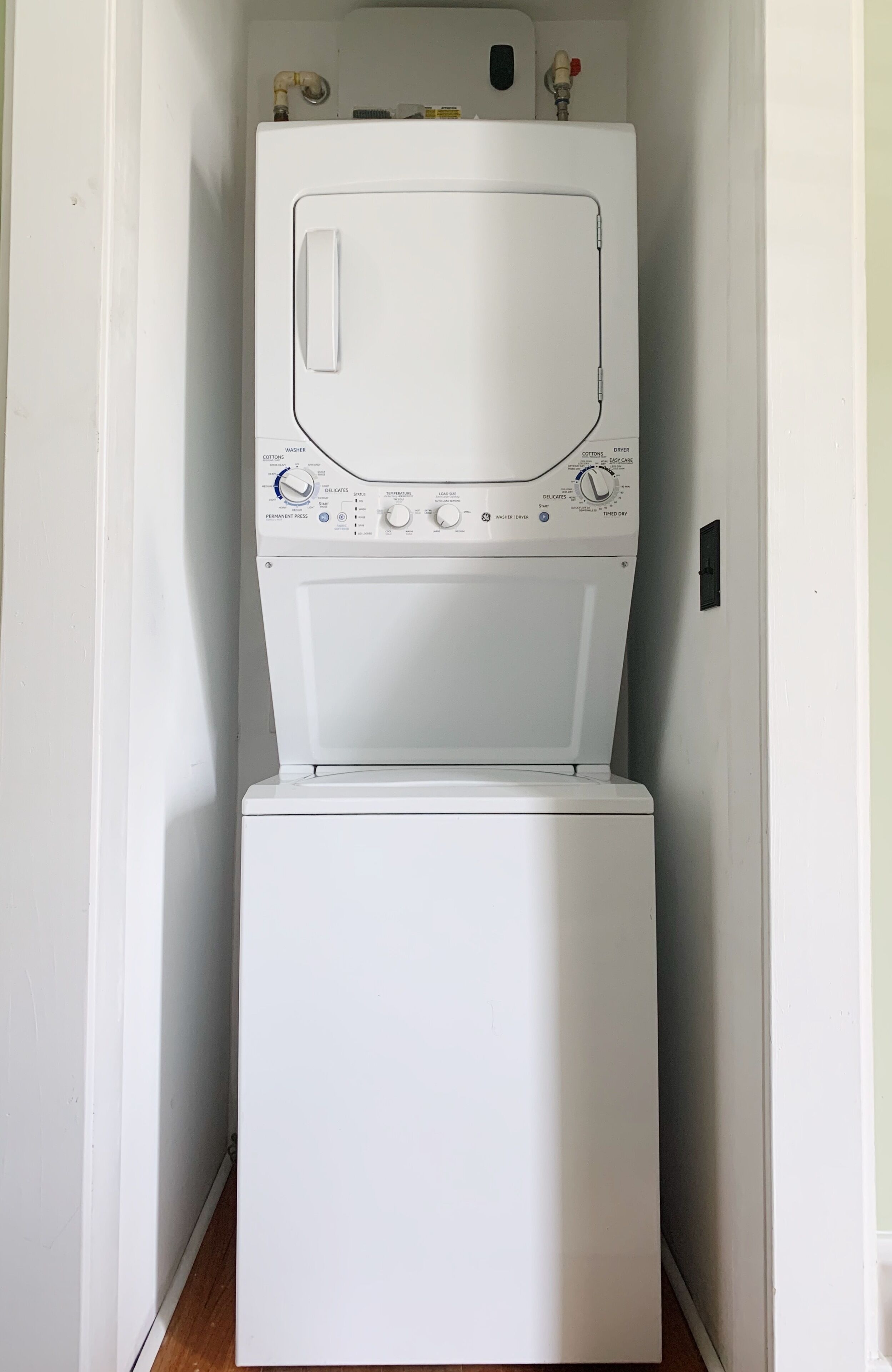 A compact, white front-loading washer and dryer unit in a narrow hallway with white walls and wooden flooring.