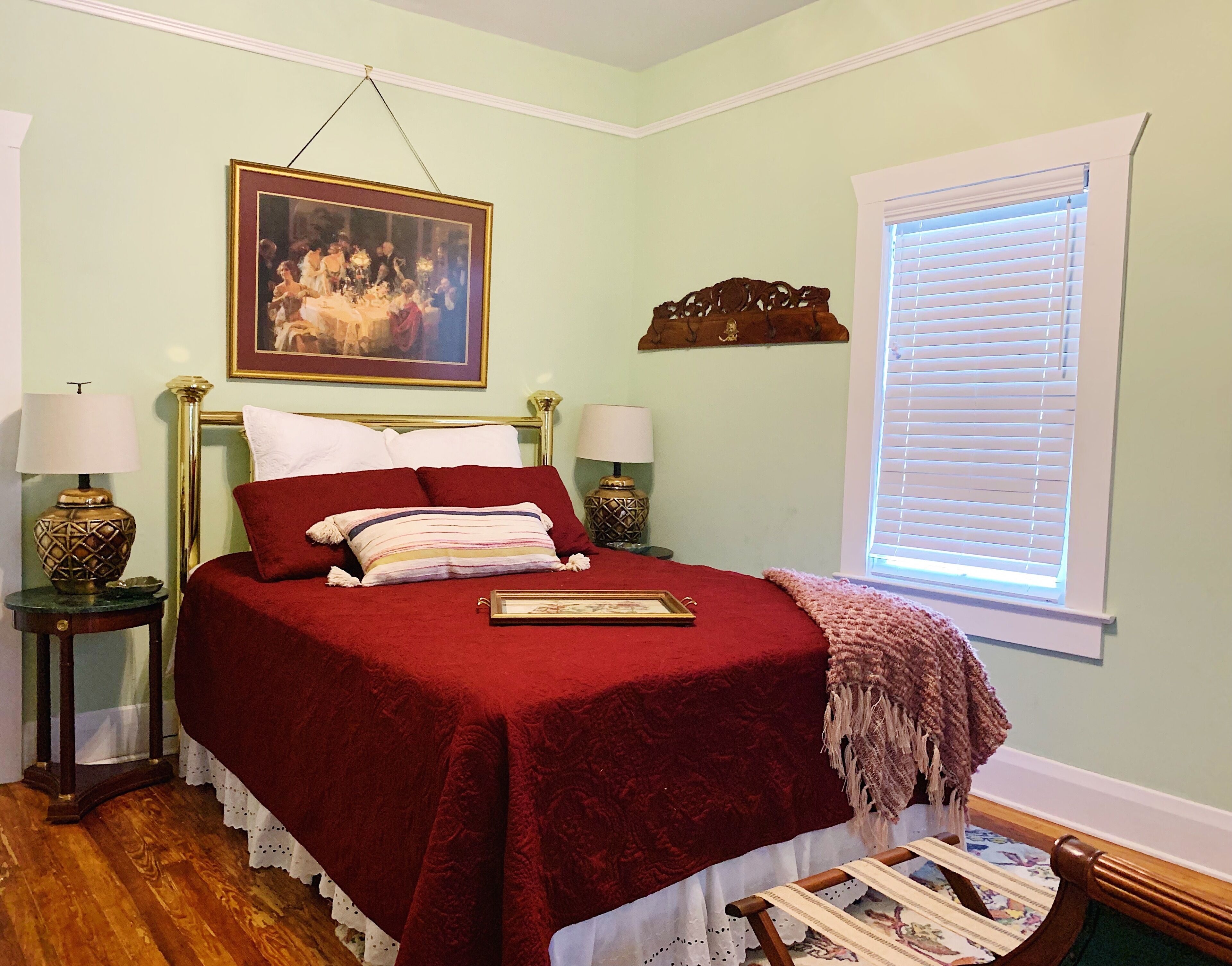 A bedroom with a bed, two bedside tables, a painting, a window with blinds, and a wooden floor.