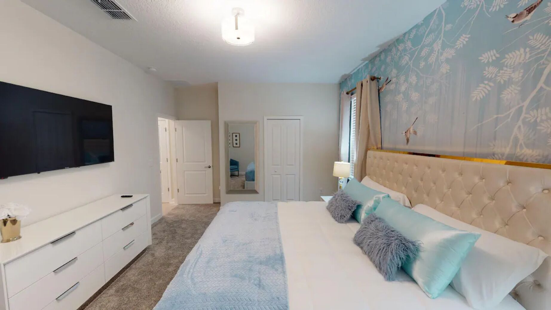 Une chambre à coucher avec un grand lit, un téléviseur à écran plat, une commode blanche et une porte donnant sur une autre pièce.