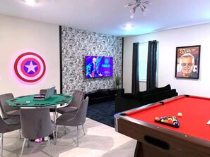 Una habitación con una mesa de billar, una mesa verde con sillas, un logotipo de Capitán América, un televisor en la pared y un cartel.