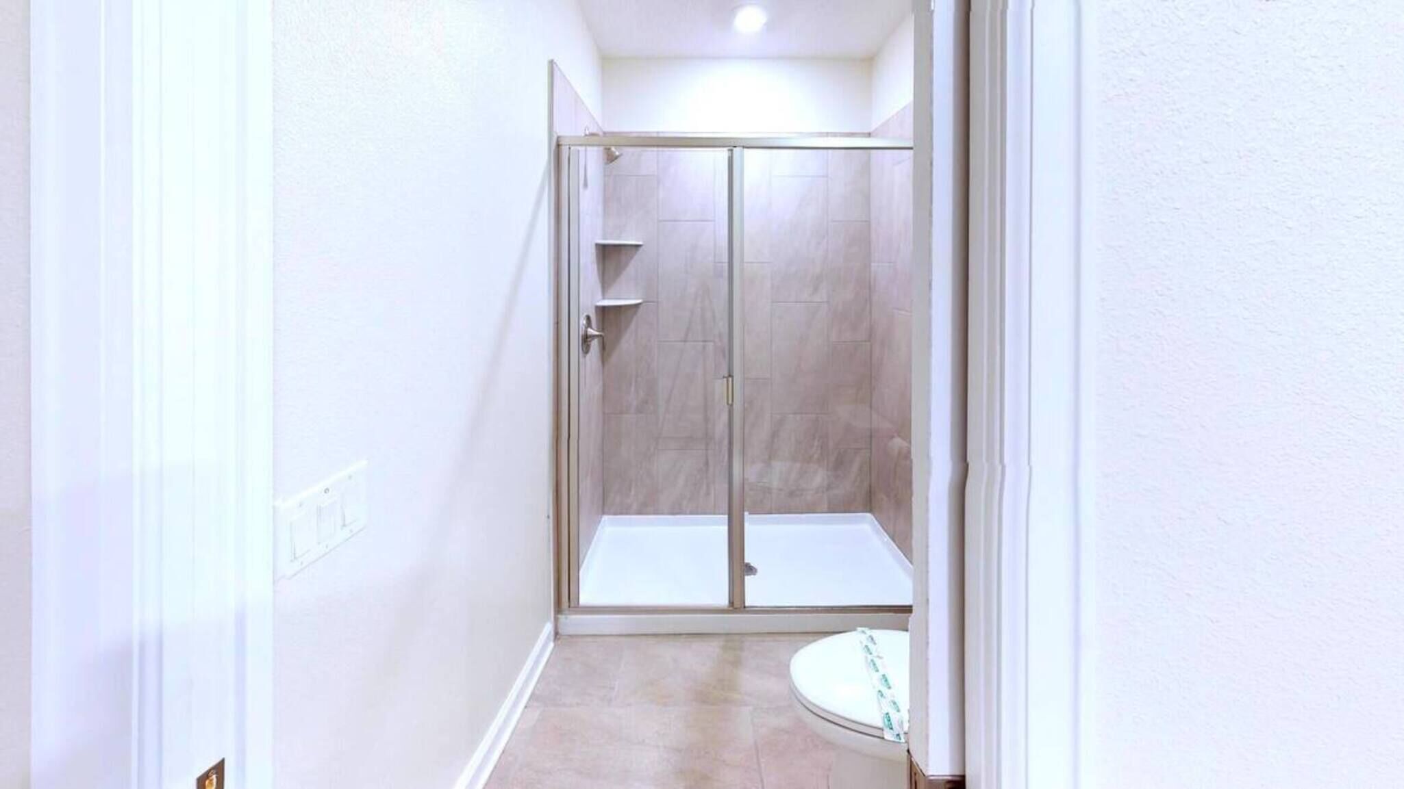 Un couloir étroit mène à un espace douche doté d’une porte en verre, d’une petite étagère et de toilettes visibles.