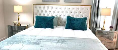 A bedroom with a tufted headboard, two teal pillows, a white bedspread, and two wall-mounted abstract paintings.