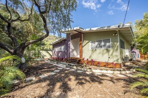 A single-story house with a porch, surrounded by a garden with various plants and a large tree. - Papillon Bleu (Horseshoe Bay)