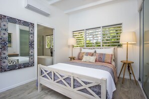 A bedroom with a bed, bedside lamps, a large mirror, and a window with blinds. - Papillon Bleu (Horseshoe Bay)