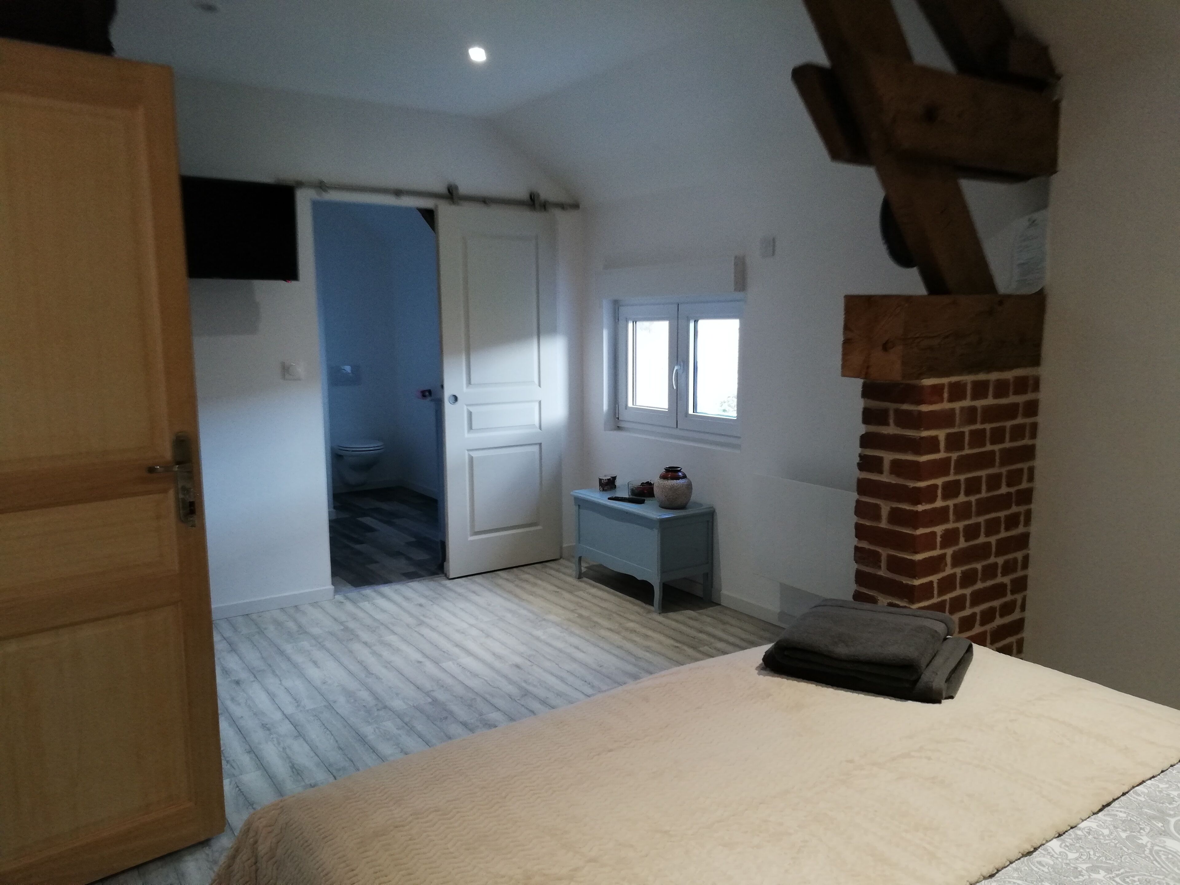 A bedroom with a bed, a small bedside table, a window, a door, and a brick wall.