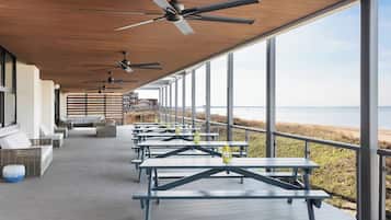 A covered outdoor area with multiple tables and chairs, large windows offering a view of the beach, and ceiling fans.