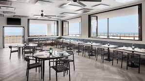 Un restaurante con mesas altas, sillas, ventanas amplias con vistas al océano, ventiladores de techo y obras de arte en las paredes.