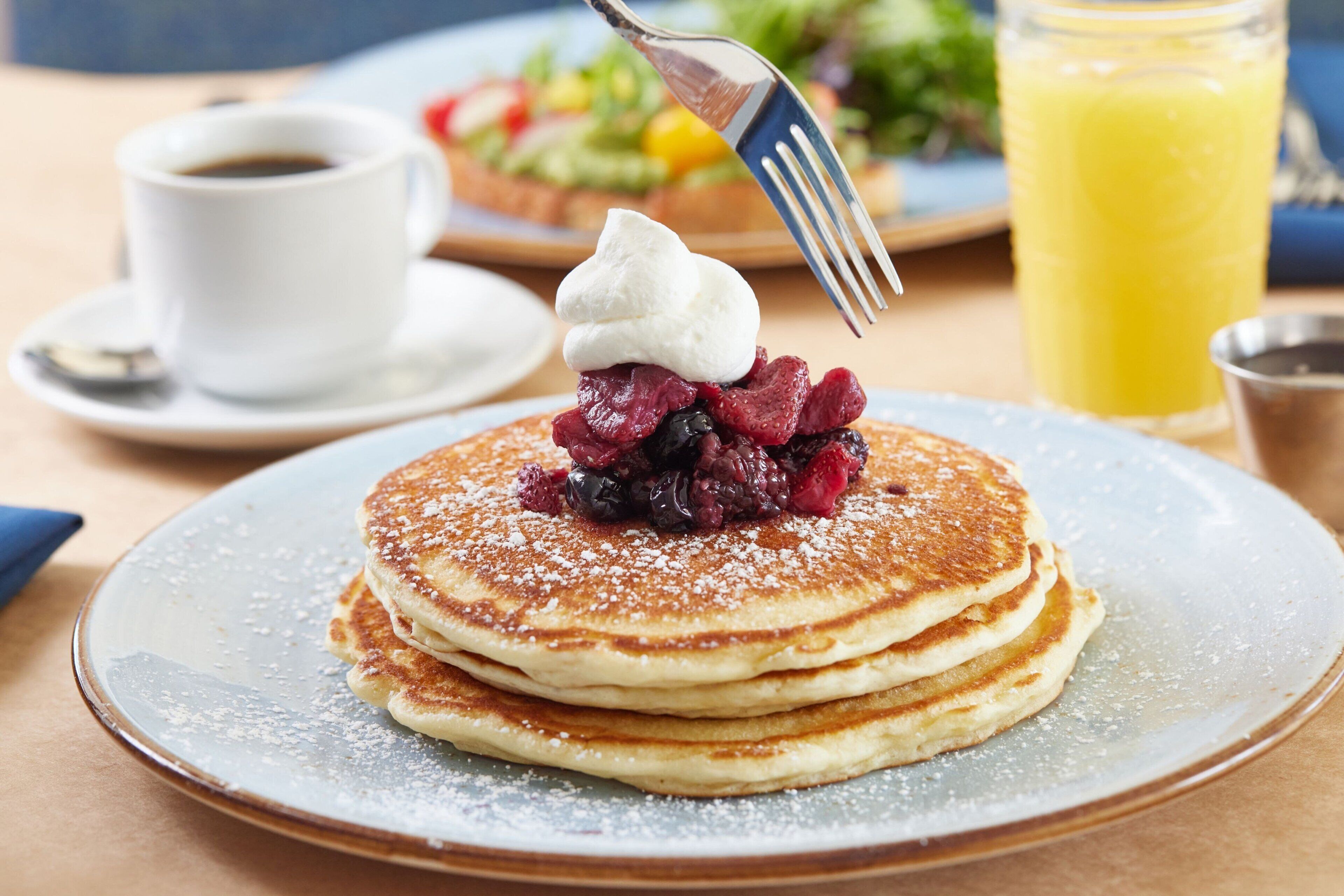 A stack of pancakes with berries and whipped cream on a plate, a cup of coffee, and a glass of orange juice on the table.
