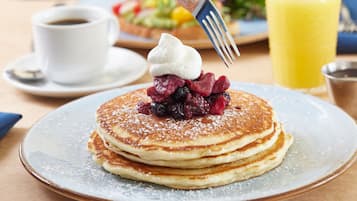 A stack of pancakes with berries and whipped cream on a plate, a cup of coffee, and a glass of orange juice on the table.