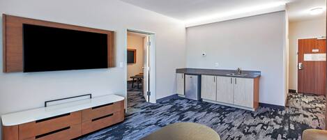 A modern hotel room with a flat-screen TV mounted on the wall, a kitchenette with a sink and cabinets, a dining area with a table and chairs, and a patterned carpet.
