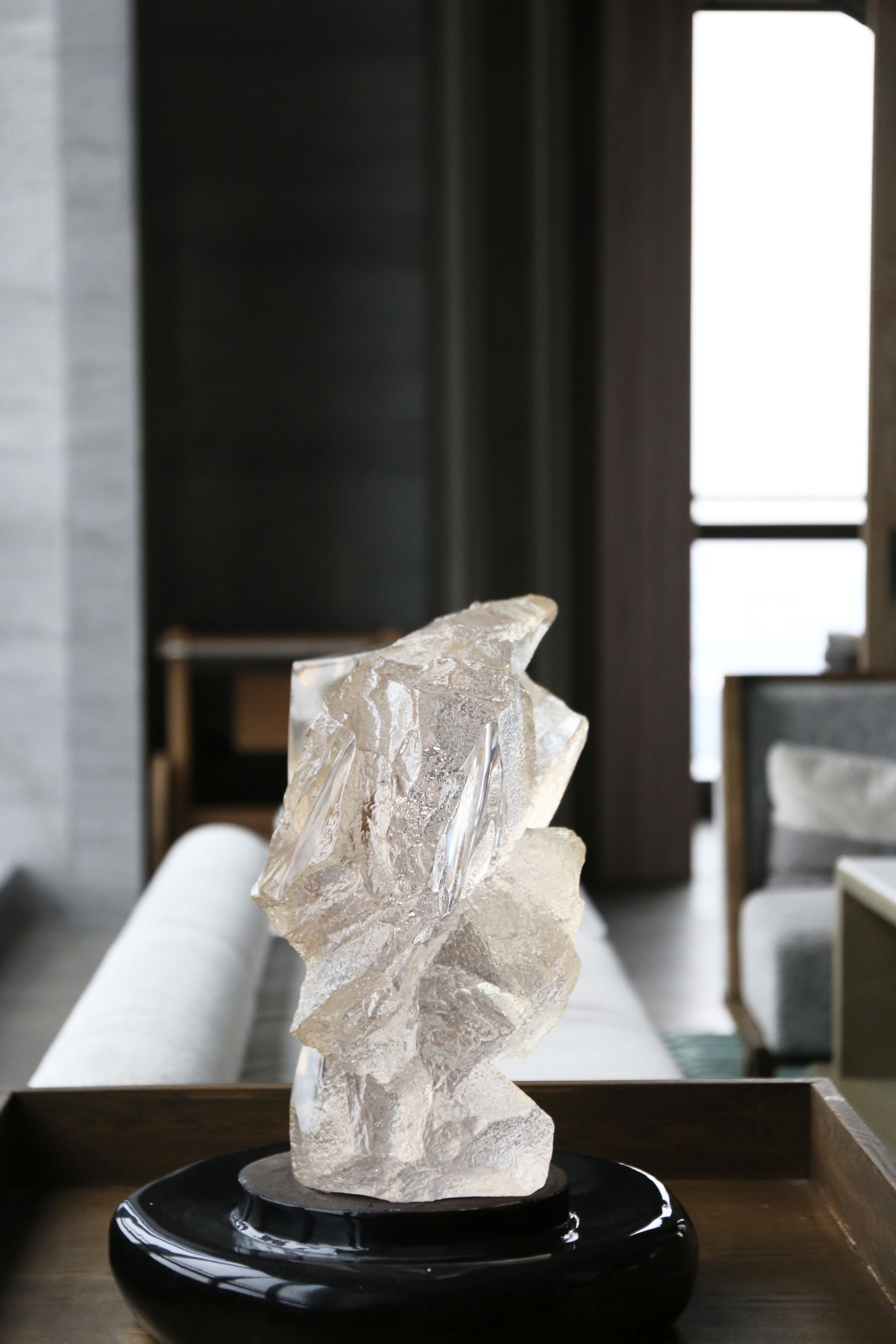 Una escultura de cuarzo transparente sobre un pedestal negro en un salón moderno con un sofá y una ventana al fondo.