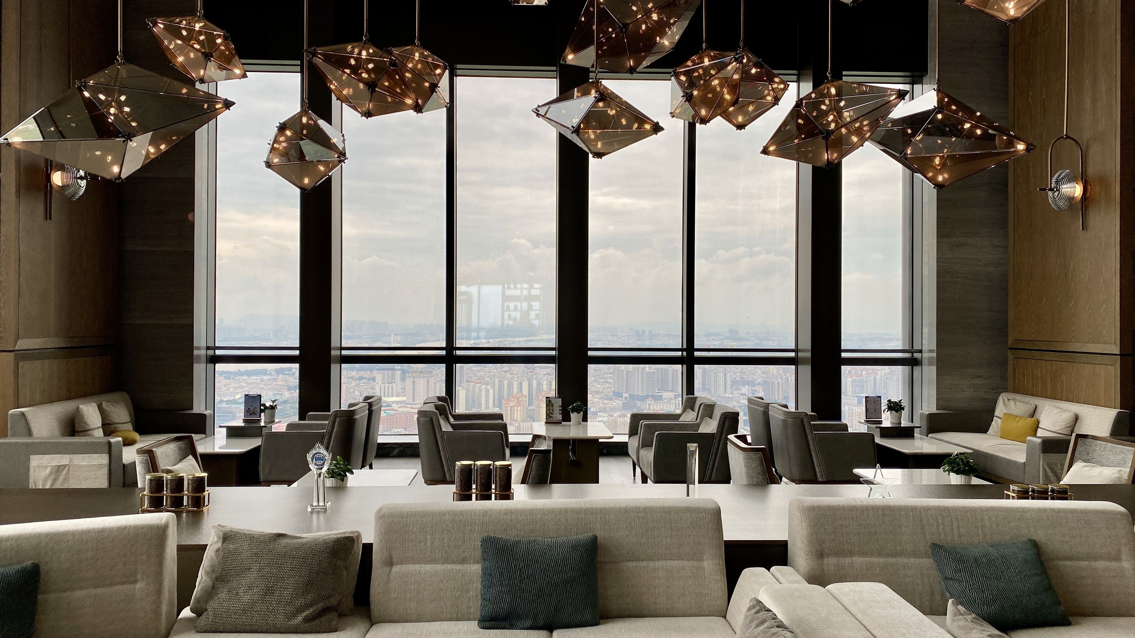 Un moderno vestíbulo de hotel con techos altos, ventanas amplias que ofrecen vistas a la ciudad y lámparas colgantes de diseño geométrico.