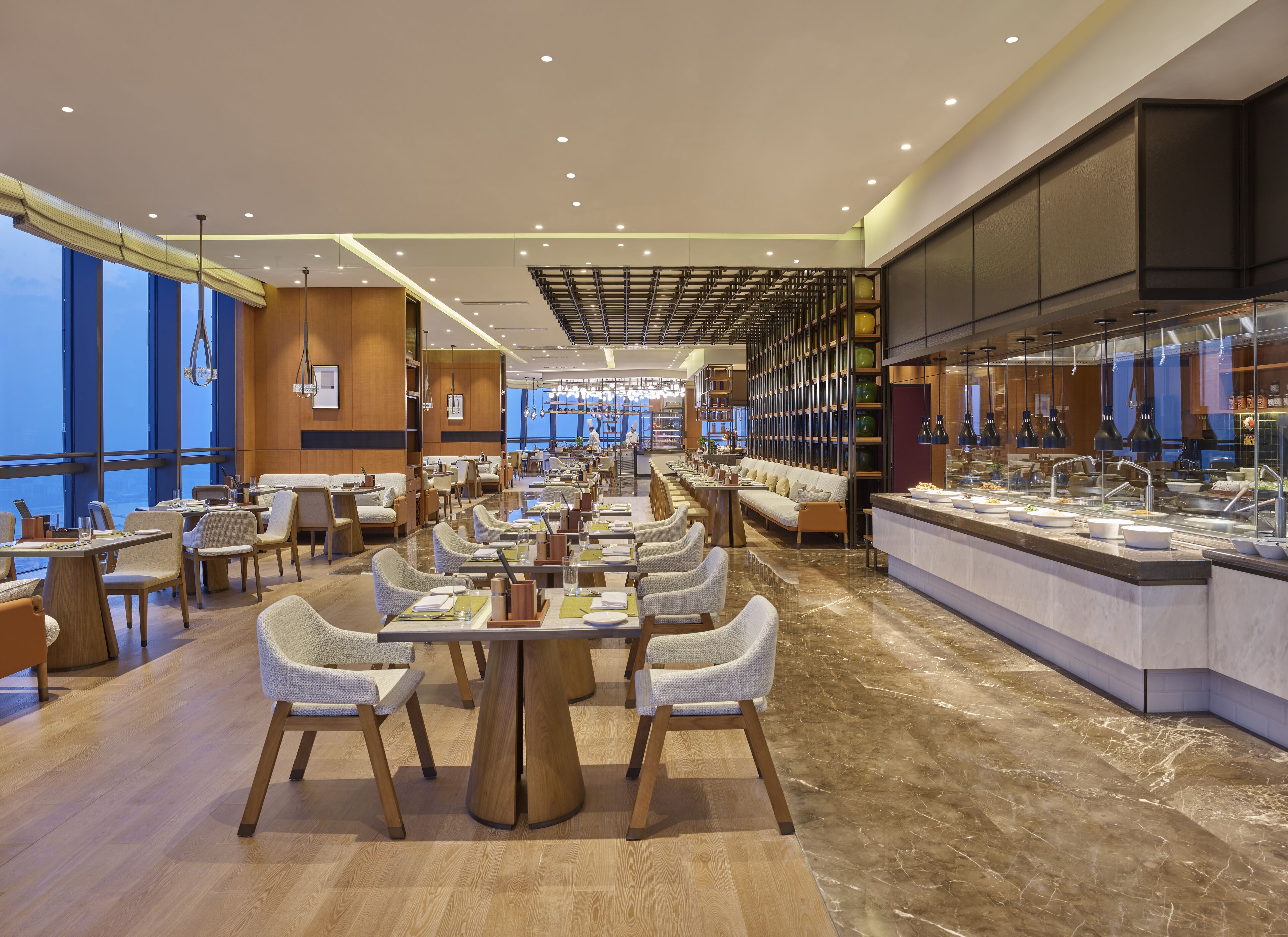 Un restaurante moderno con una amplia zona de comedor, mesas y sillas de madera. Hay un mostrador de buffet a la derecha con diferentes opciones de comida.