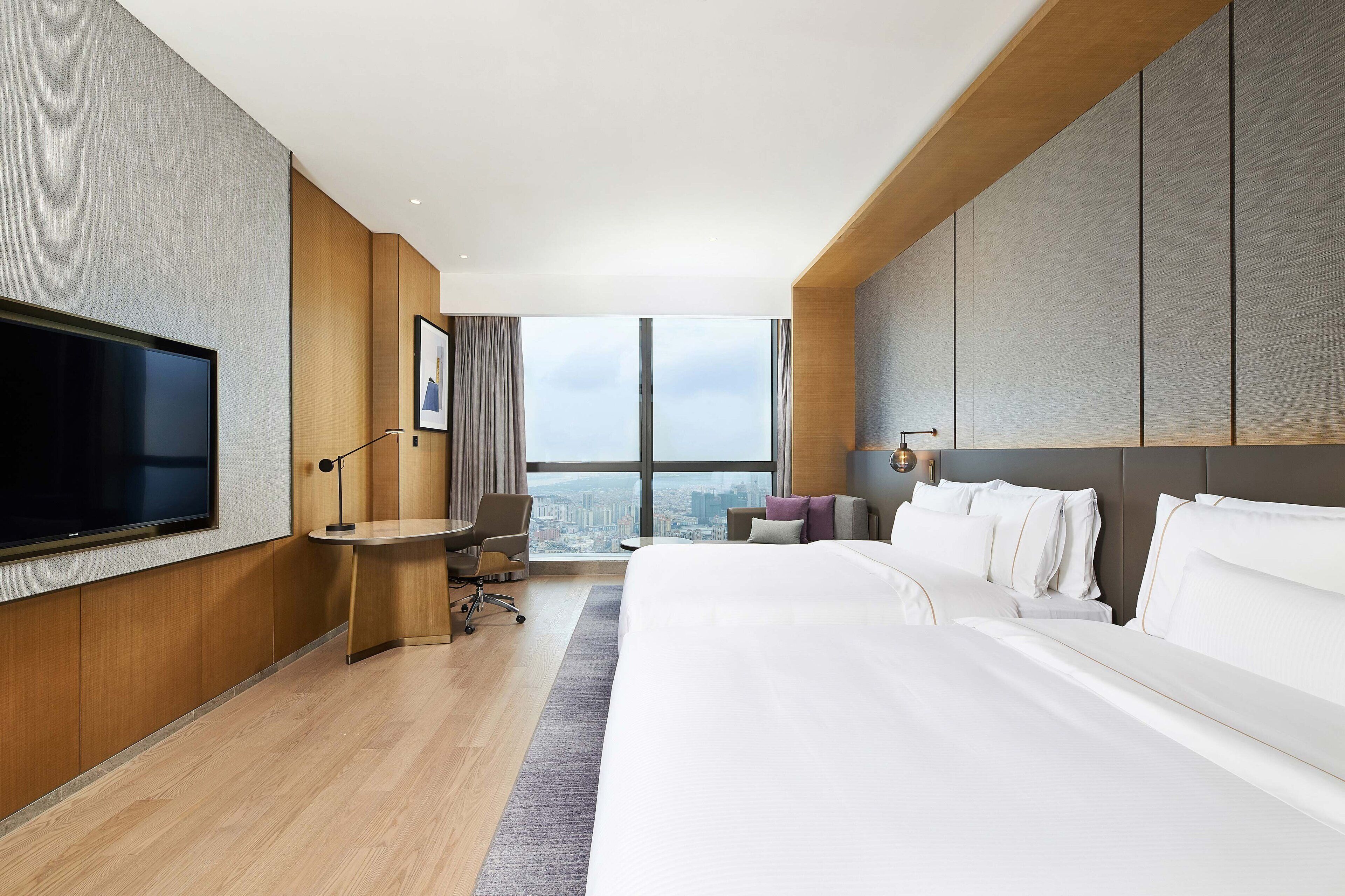 A modern hotel room with a large bed, a desk, a chair, a flat-screen TV, and a city view.