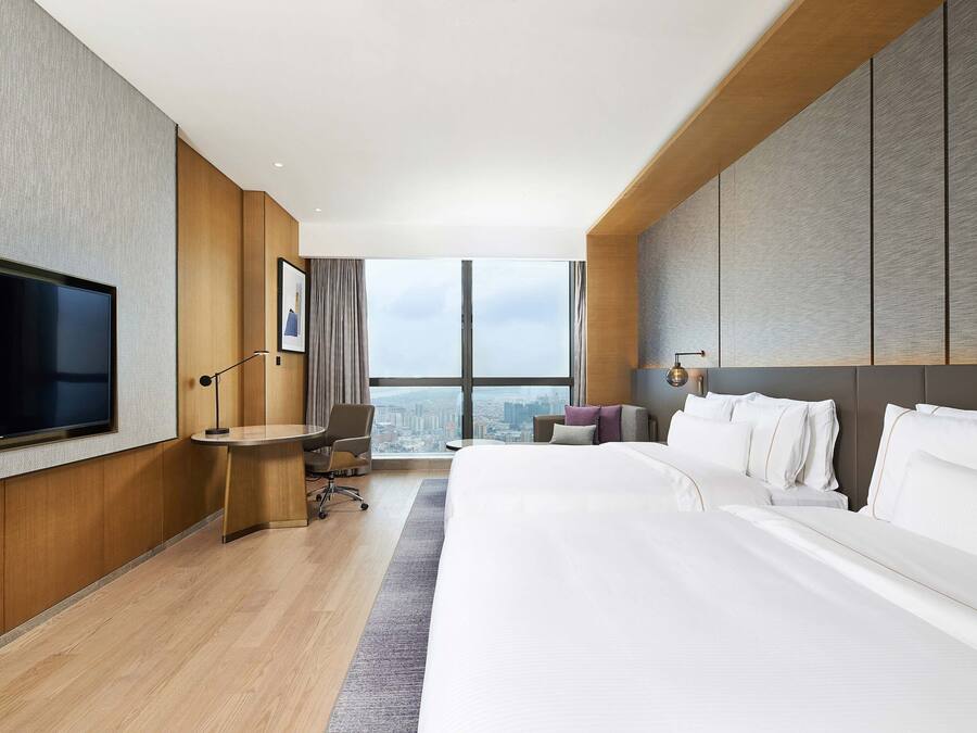 A modern hotel room with a large bed, a desk, a chair, a flat-screen TV, and a city view.