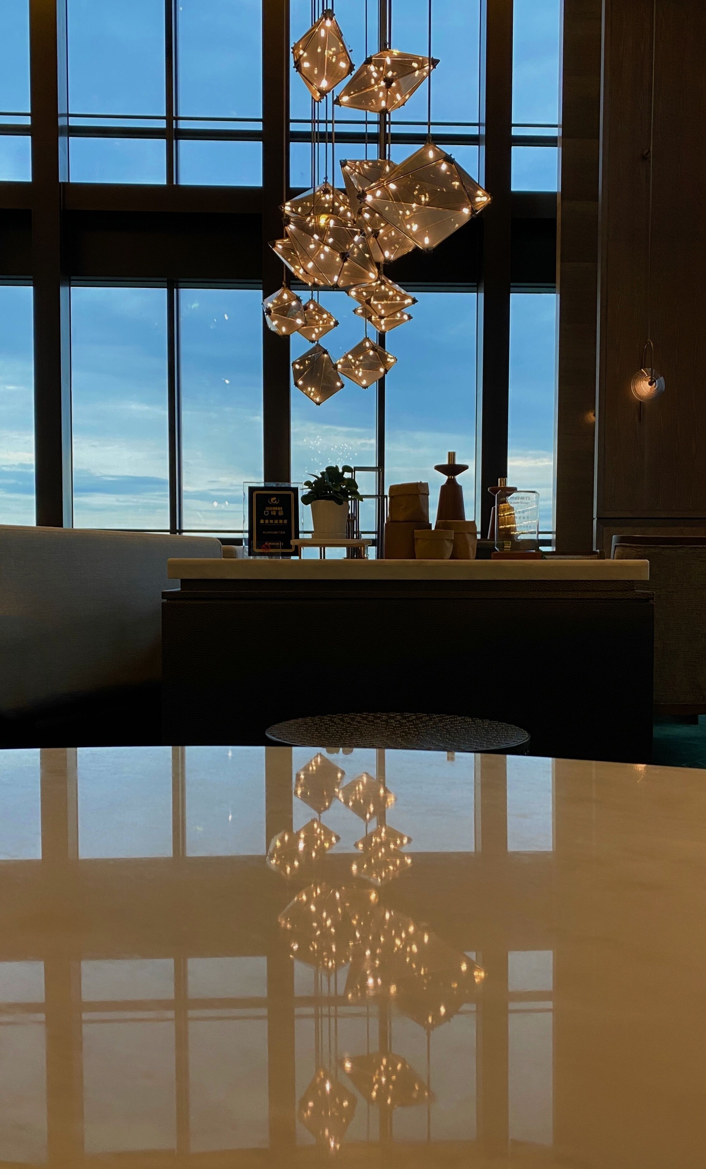 A modern interior with a reflective floor, large windows, and a geometric chandelier.