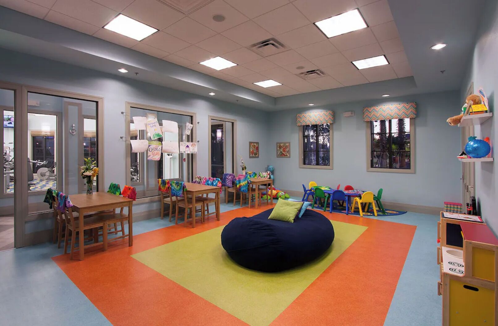 Una sala de juegos infantiles con mesas, sillas, un sillón de peluche y decoraciones coloridas.