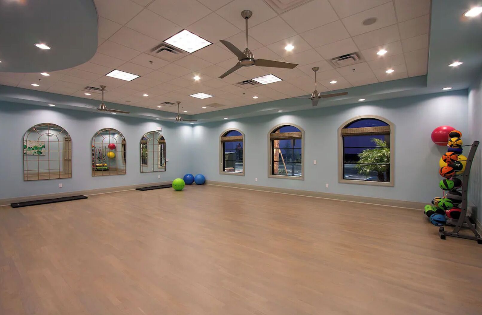 A spacious room with a wooden floor, arched windows, and ceiling fans.