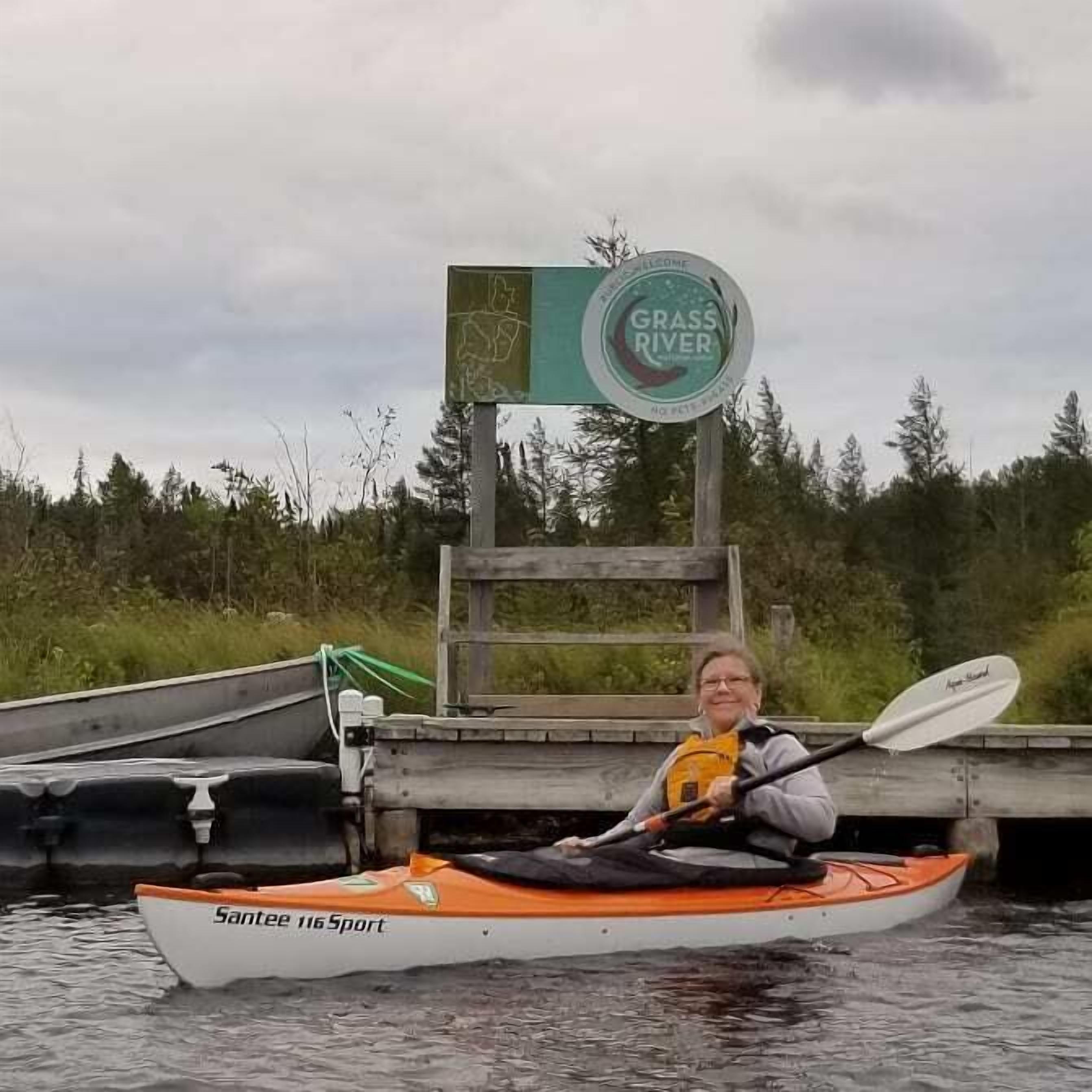 A person in a canoe on a body of water with a sign that reads "GRASS RIVER" in the background.
