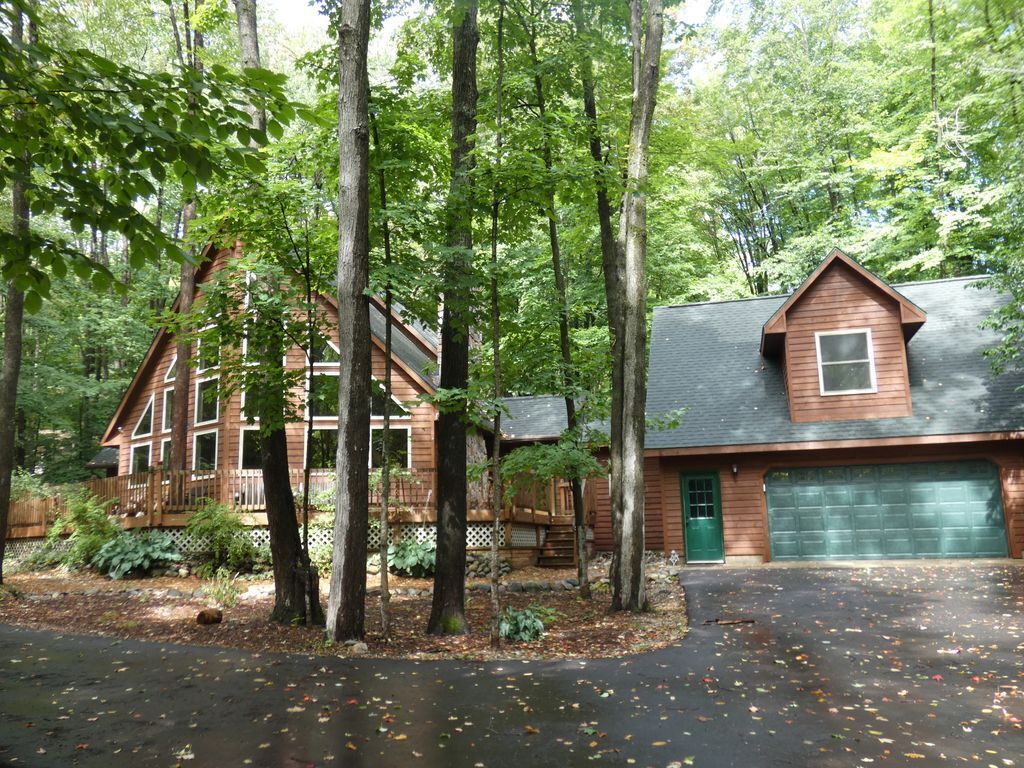 Une cabane avec un garage, entourée de grands arbres et dotée d’une terrasse en bois.