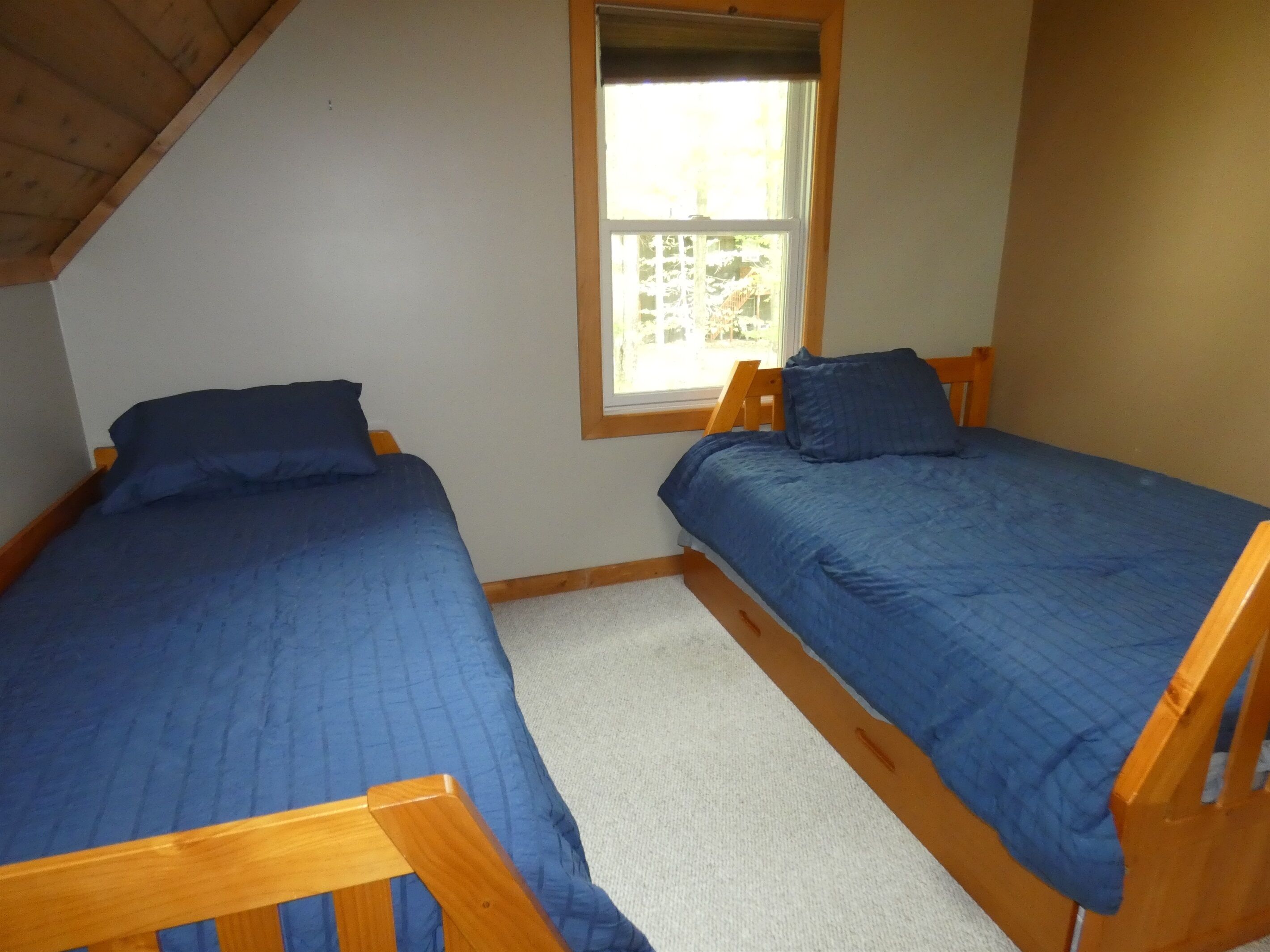 A small room with two wooden beds, each with a blue mattress and matching pillows. There is a window with a wooden frame, and the room has white walls and a sloped ceiling.