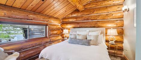 Een slaapkamer in een blokhut met een groot bed, twee bedlampen en een raam met uitzicht op een natuurlandschap.