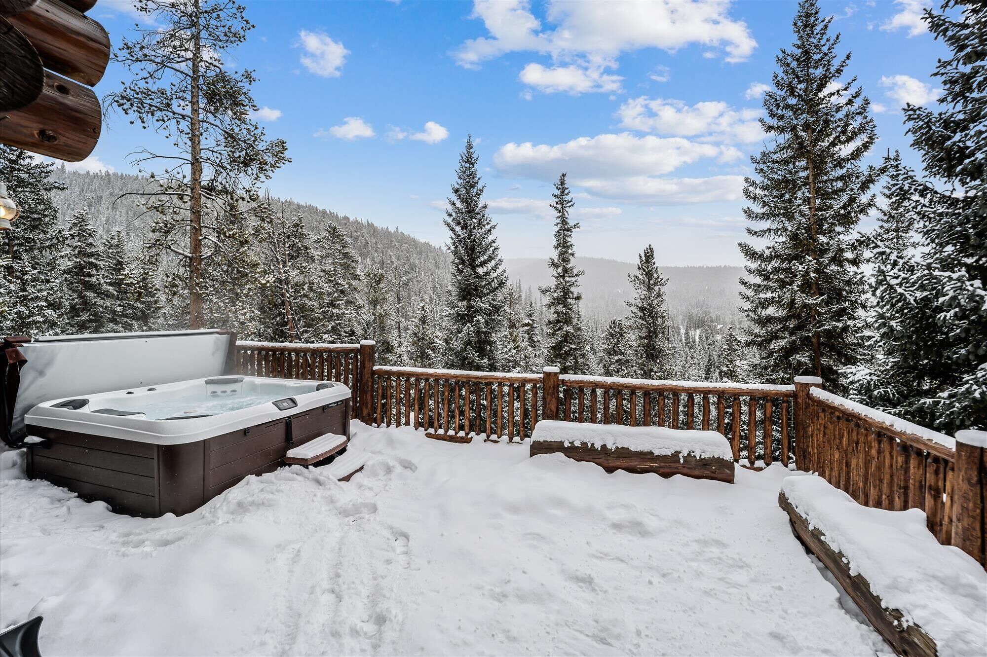 A hot tub on a wooden deck surrounded by snow-covered trees and a mountainous backdrop.
