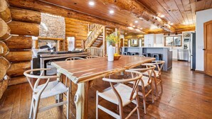 A log cabin interior with a wooden dining table, chairs, a stone fireplace, and a kitchen area. - Baldy Mtn Log Ski Home, Game Room, Free Shuttle, Sleeps 12 (Breckenridge)