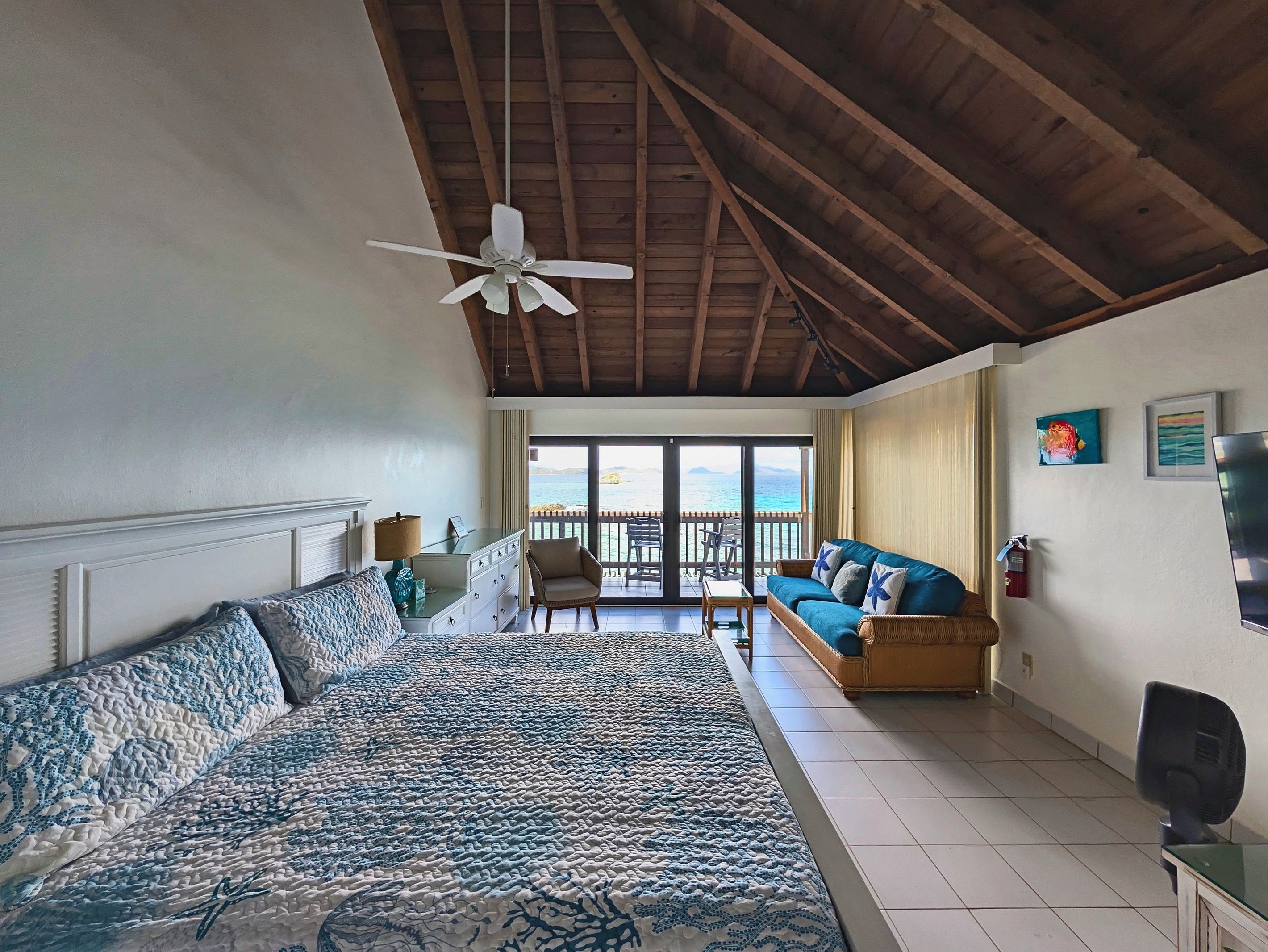 A bedroom with a large bed, a ceiling fan, a balcony with a view of the sea, and a sofa.