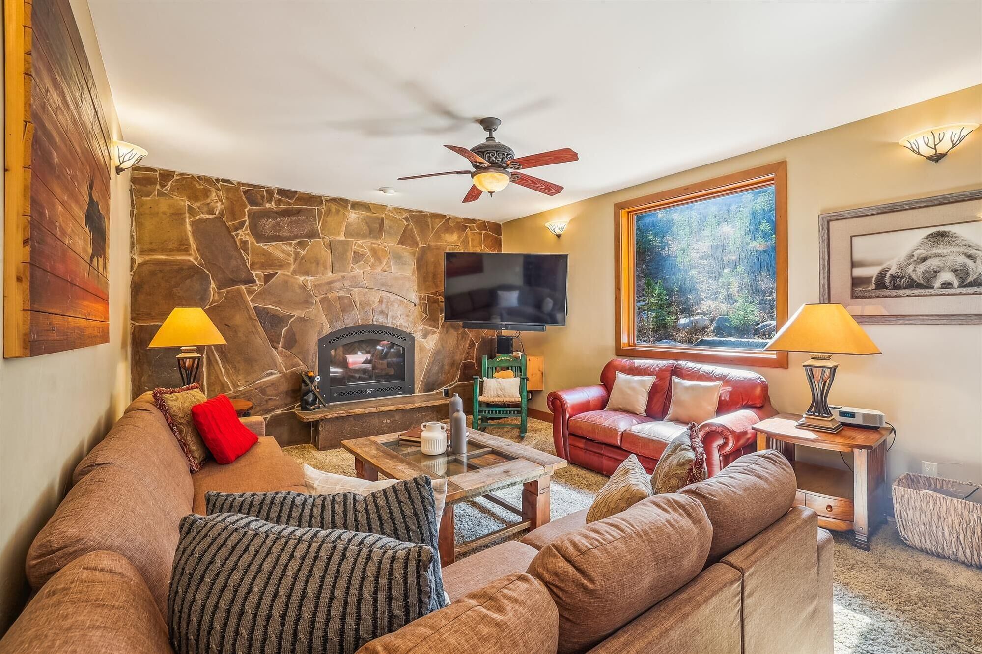 A cozy living room with a stone fireplace, leather sofa, and a large window offering a forest view.