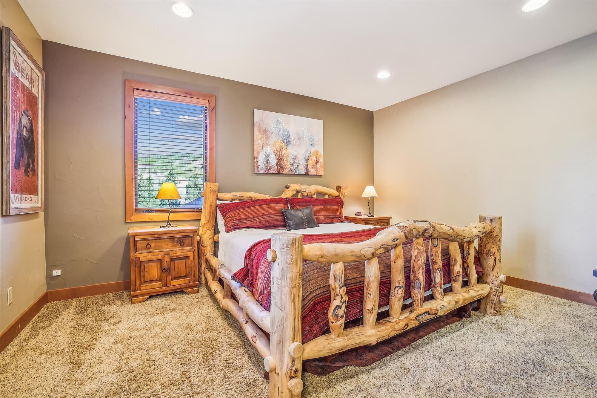 A bedroom with a wooden bed frame, two bedside tables, a window with blinds, and artwork on the walls.