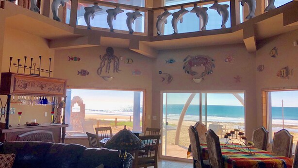 Ein geräumiger Wohnbereich mit Blick auf den Strand, ein Essbereich mit Tisch und Stühlen sowie ein Balkon mit Delfin-Skulpturen.