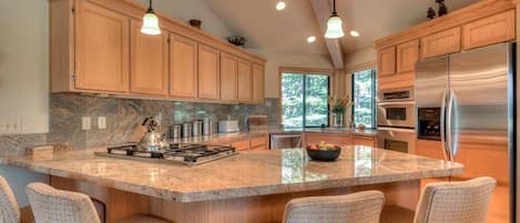 A spacious kitchen with wooden cabinets, a large island with bar stools, stainless steel appliances, and a view of the outdoors through the windows.