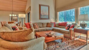 A spacious living room with a large sectional sofa, a coffee table, and a patterned rug. There are multiple windows providing natural light and a view of the outdoors. - Luxurious home on the golf course (Sunriver)