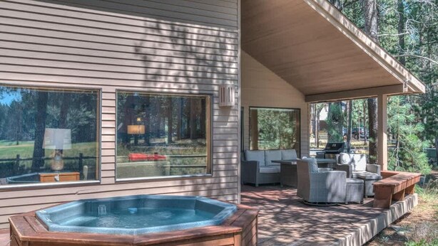 A wooden hot tub in a covered outdoor area with a deck, seating, and large windows overlooking a forested area. - Luxurious home on the golf course (Sunriver)
