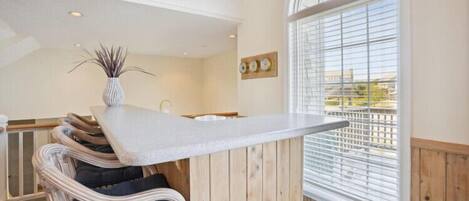 Una habitación con techo alto, una mesa de comedor con sillas, una ventana con persianas y vistas al exterior.