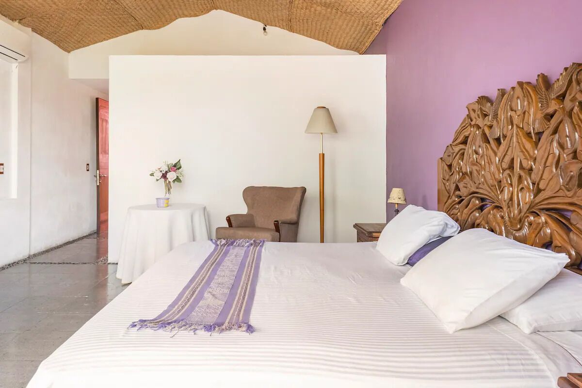 Une chambre d’hôtel avec un grand lit, une tête de lit en bois, une chaise, une petite table sur laquelle est posé un vase de fleurs et une lampe.