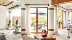 Amplio espacio interior con un gran ventanal corredero que da a un patio exterior. Hay varias sillas y una mesa con un jarrón de flores.