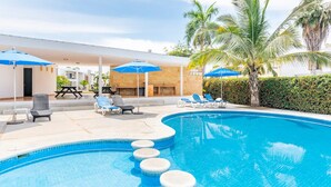 Una piscina con sillas de descanso y sombrillas, rodeada de palmeras y una zona de estar techada.