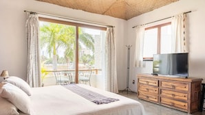 Un dormitorio con una cama grande, un televisor sobre una cómoda de madera y un balcón con vistas a palmeras.