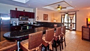 Uma cozinha moderna com armários de madeira escura, uma ilha central com banquetas e um ventilador de teto, além de uma grande janela com cortinas.